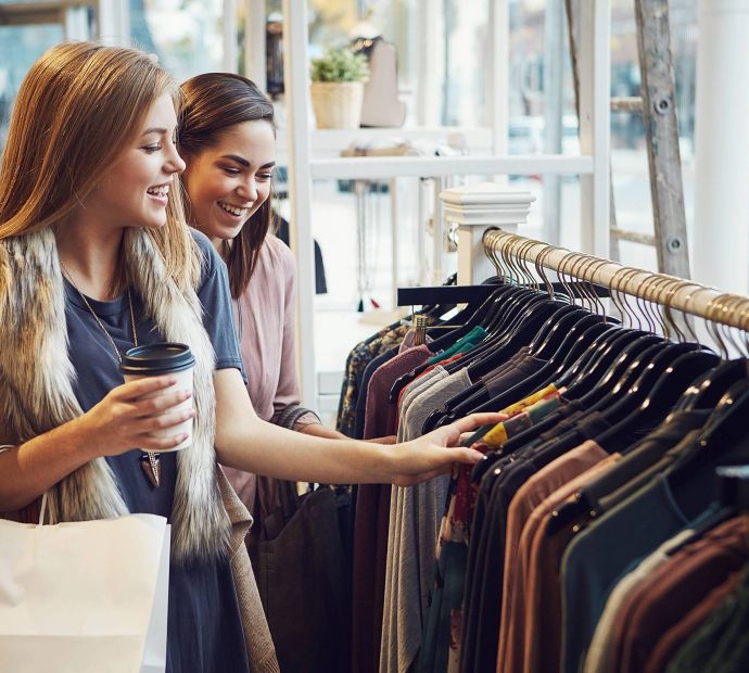 The Narrows Two young women smiling and shopping for clothes together, looking at items on a rack in a clothing store.