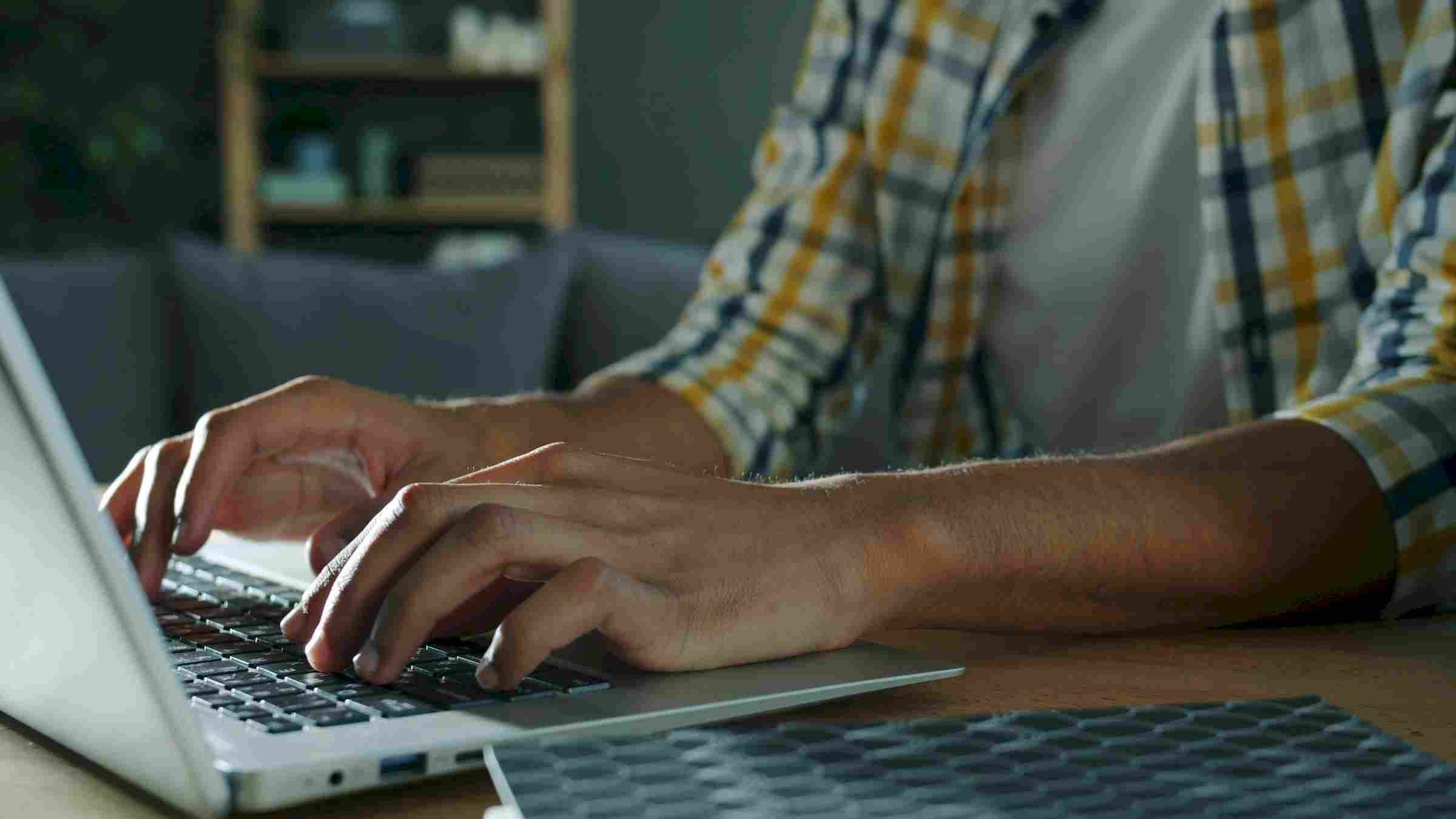The Narrows Close-up of a person typing on a laptop keyboard, wearing a plaid shirt, with a blurred background.