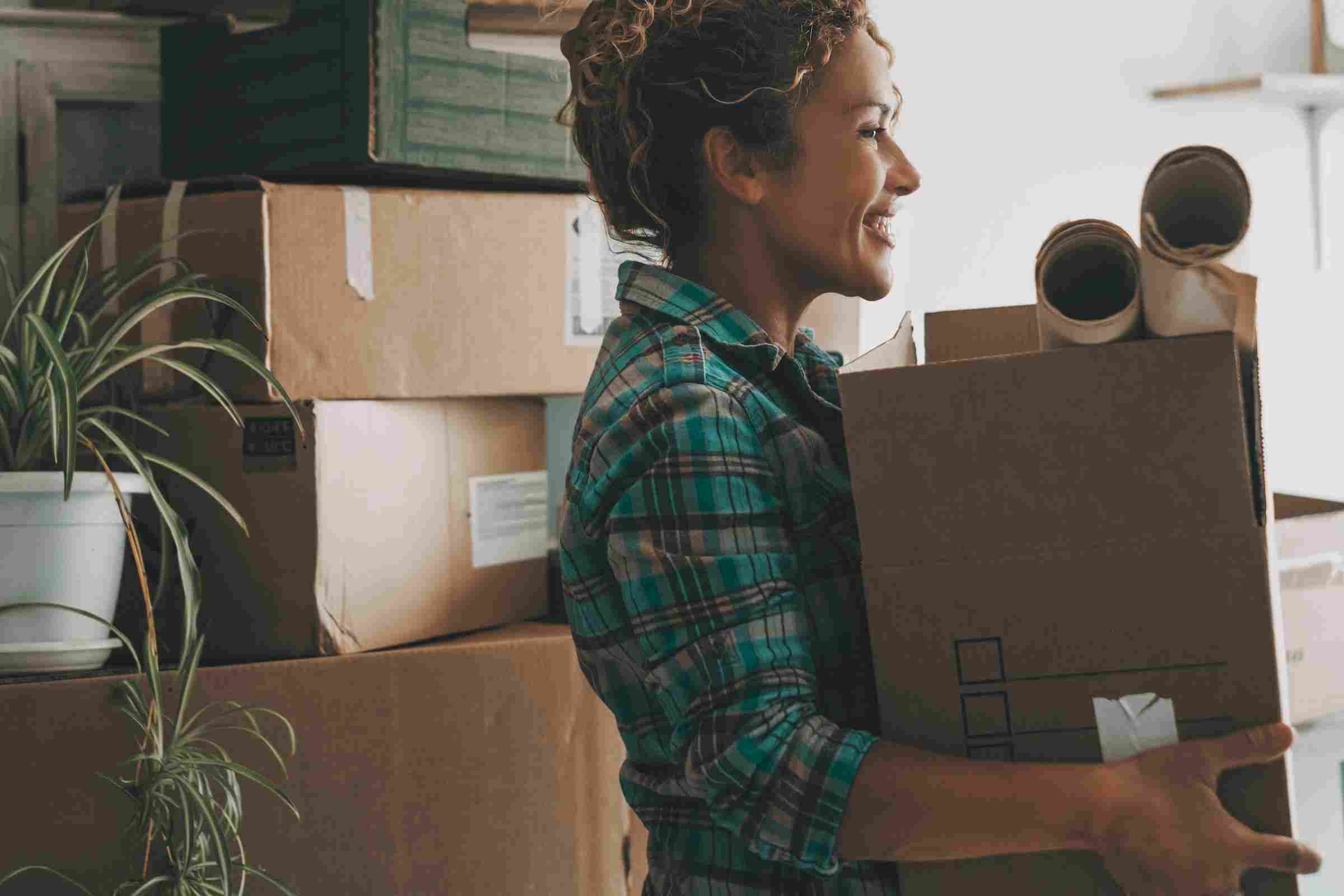 The Narrows Smiling woman carrying moving boxes with packing supplies, surrounded by more boxes and a potted plant.