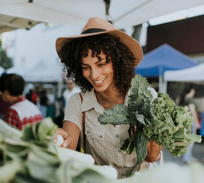 Allers Landing A woman smiles while holding leafy greens at an outdoor farmers market.