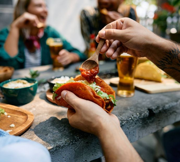 The Narrows A person adds salsa to a taco at an outdoor table with friends enjoying food and drinks.